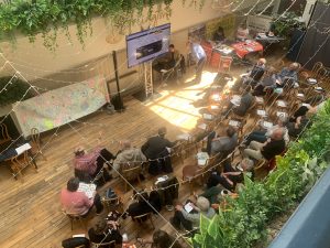 A bird's eye view of an audience sitting in rows watching a presentation. Speaker is presenting at the front on a large screen. To the side of the speaker are two campaigners at tables with posters protecting Sizewell C.